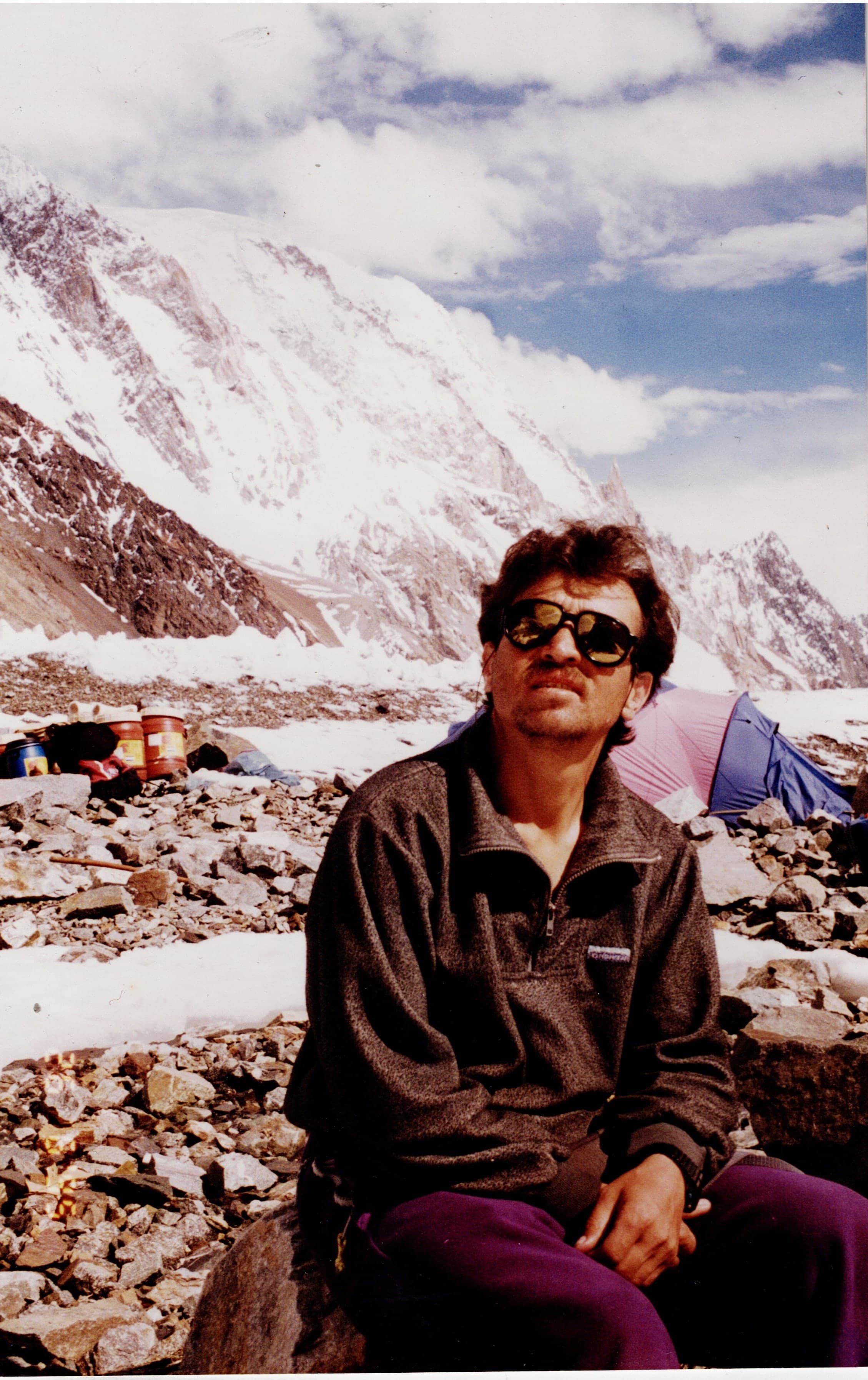 A man in sunglasses sitting on a rock at a Karakoram base camp with tents and a snow face behind