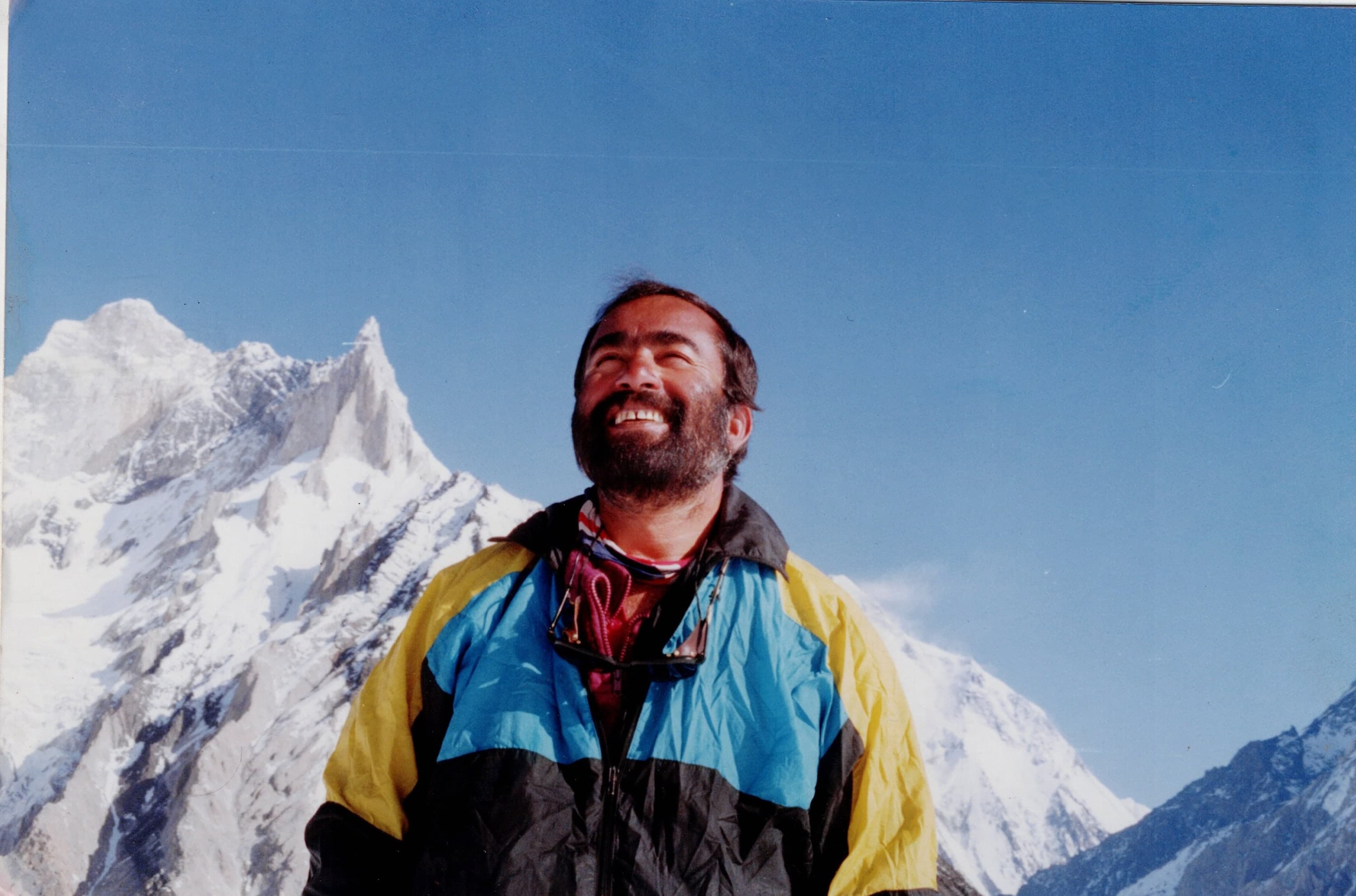 A smiling bearded man in a blue and yellow jacket below a sharp Karakoram rock spire