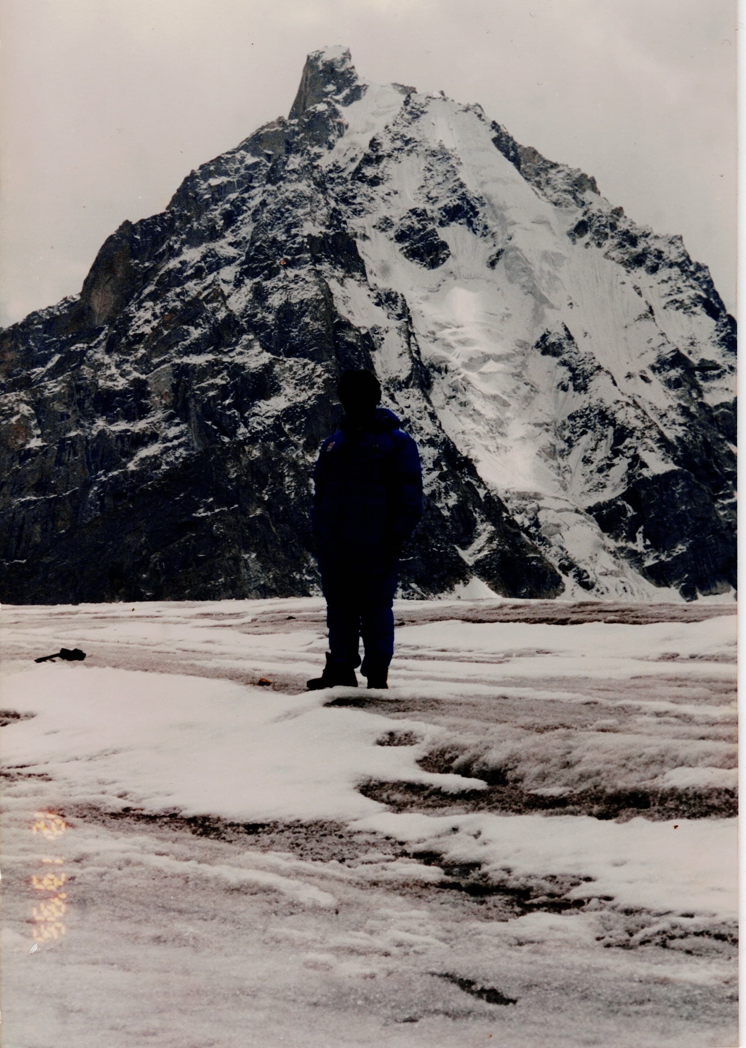 A lone figure standing on a snow-covered glacier below a steep rock and ice pyramid peak