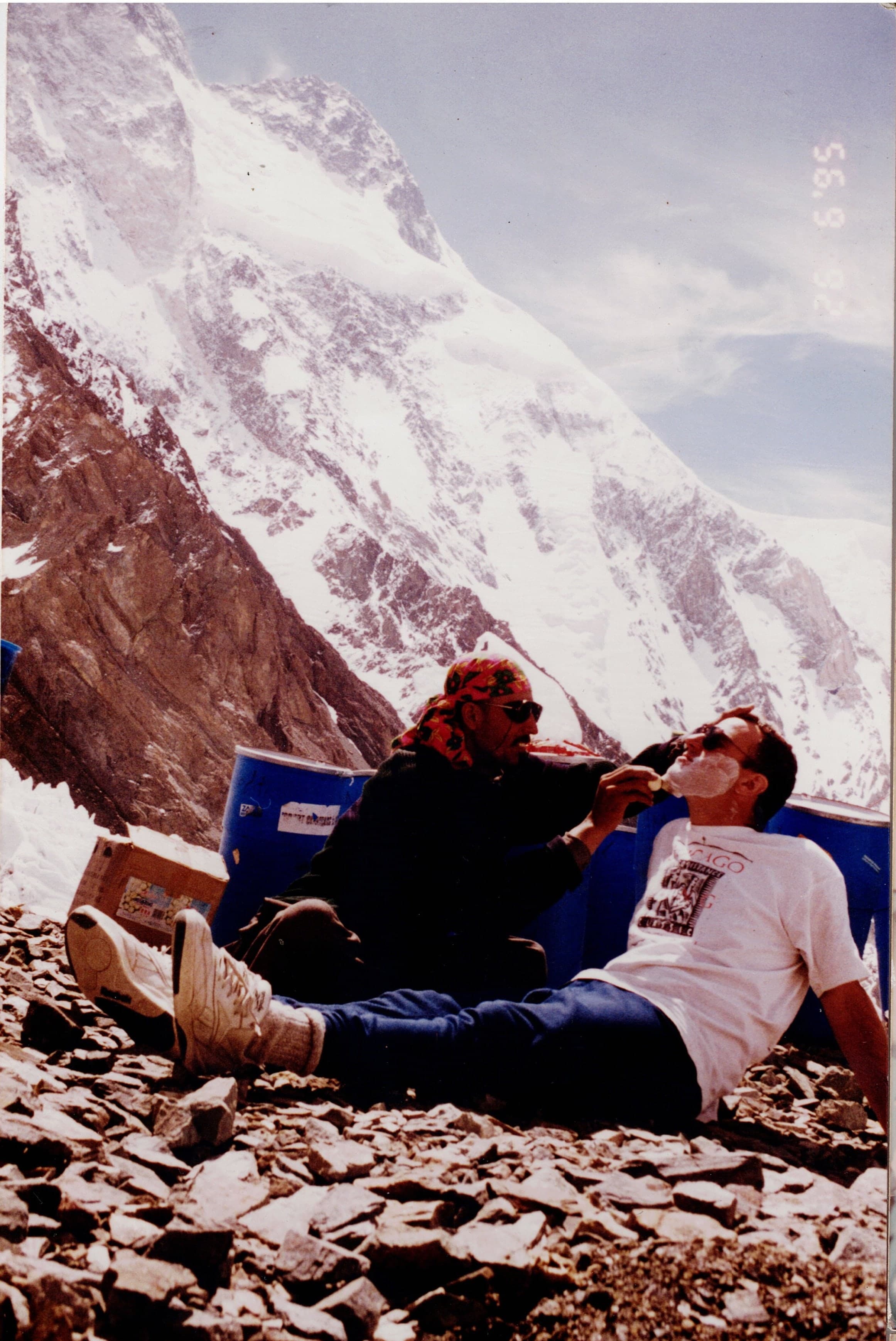 Two men joking in front of blue expedition barrels at a base camp below a huge snow face