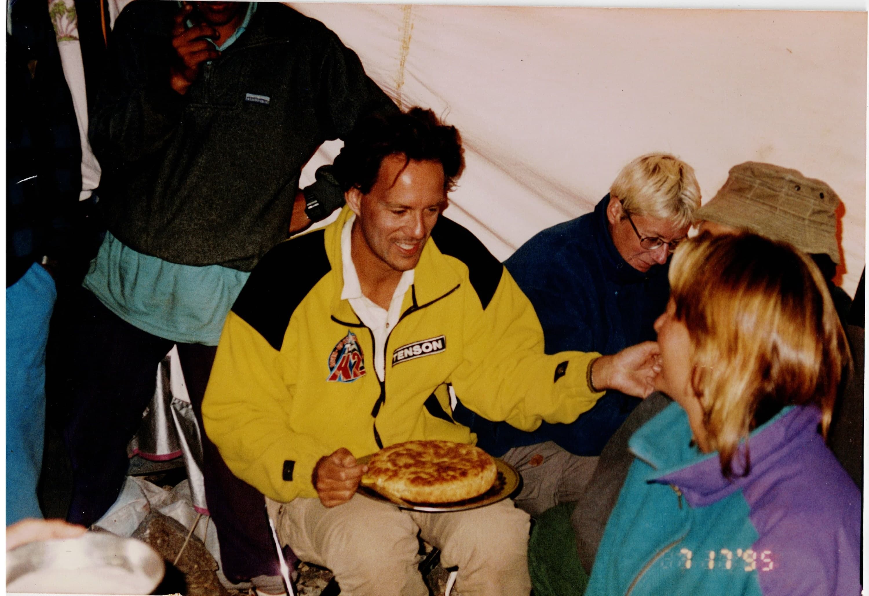 A man in a yellow jacket sharing a cake with expedition members inside a tent