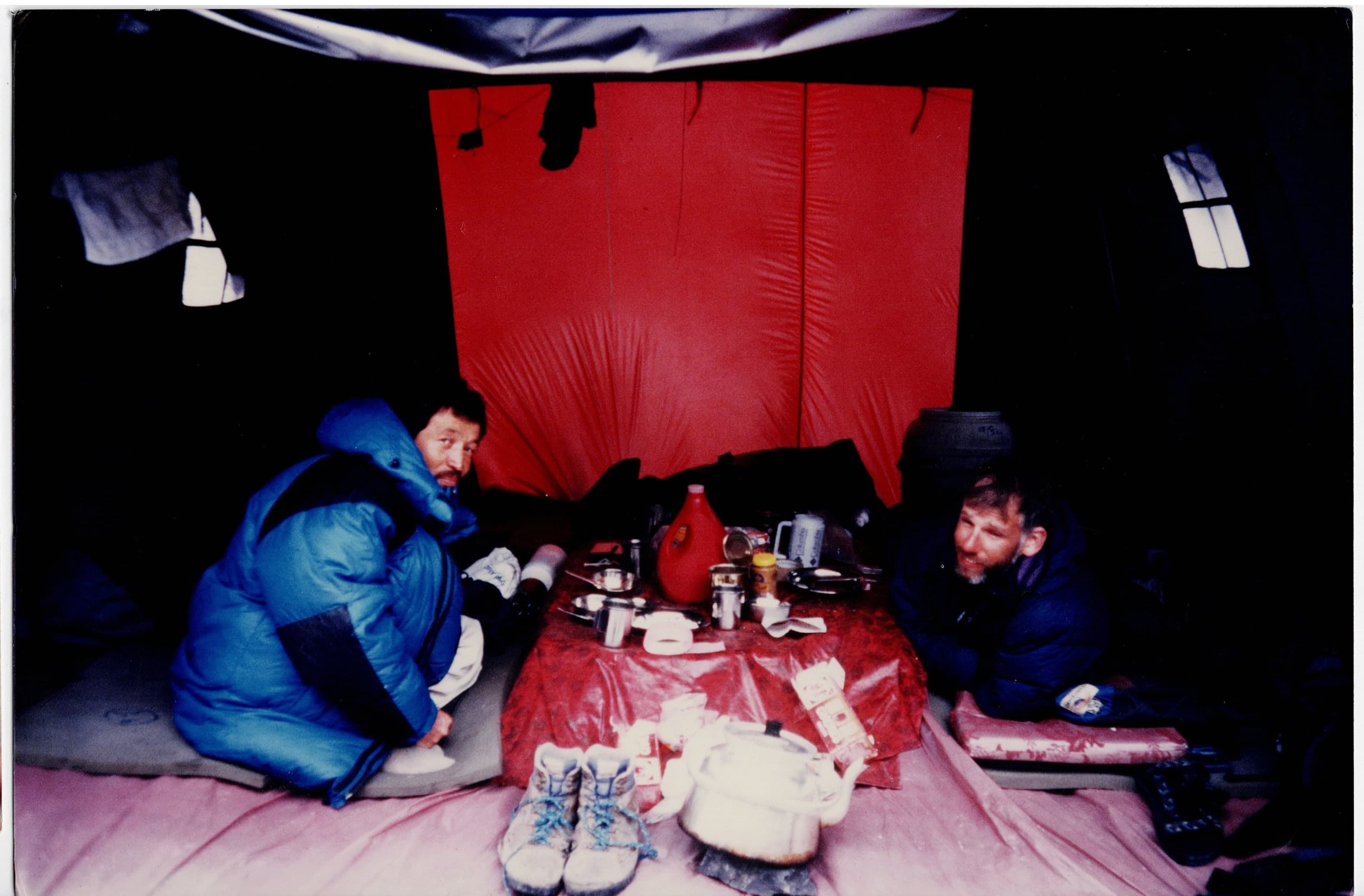 Two climbers in down jackets having tea inside a dark expedition mess tent