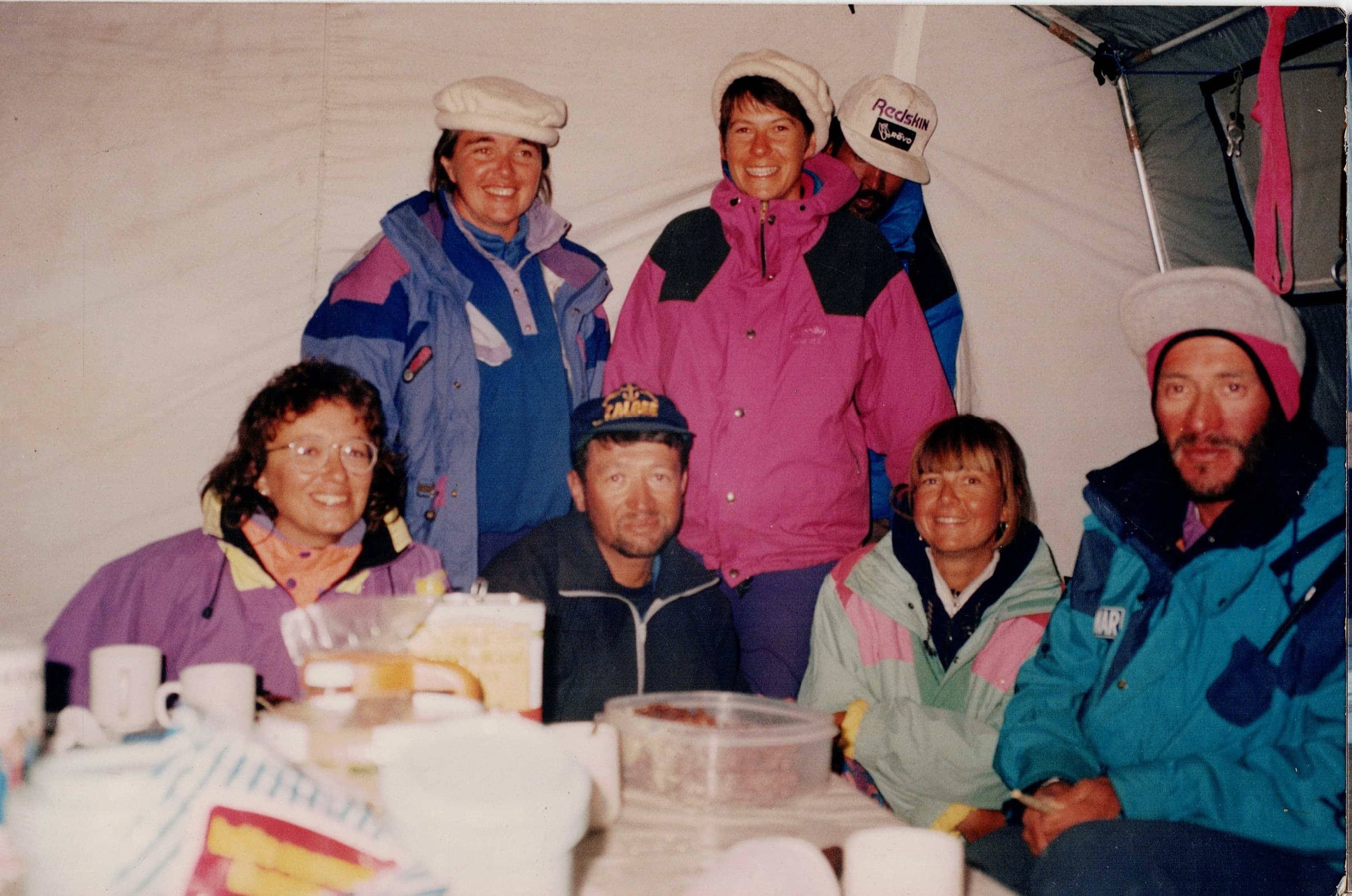 Six expedition members, several wearing white Hunza caps, around a mess tent table