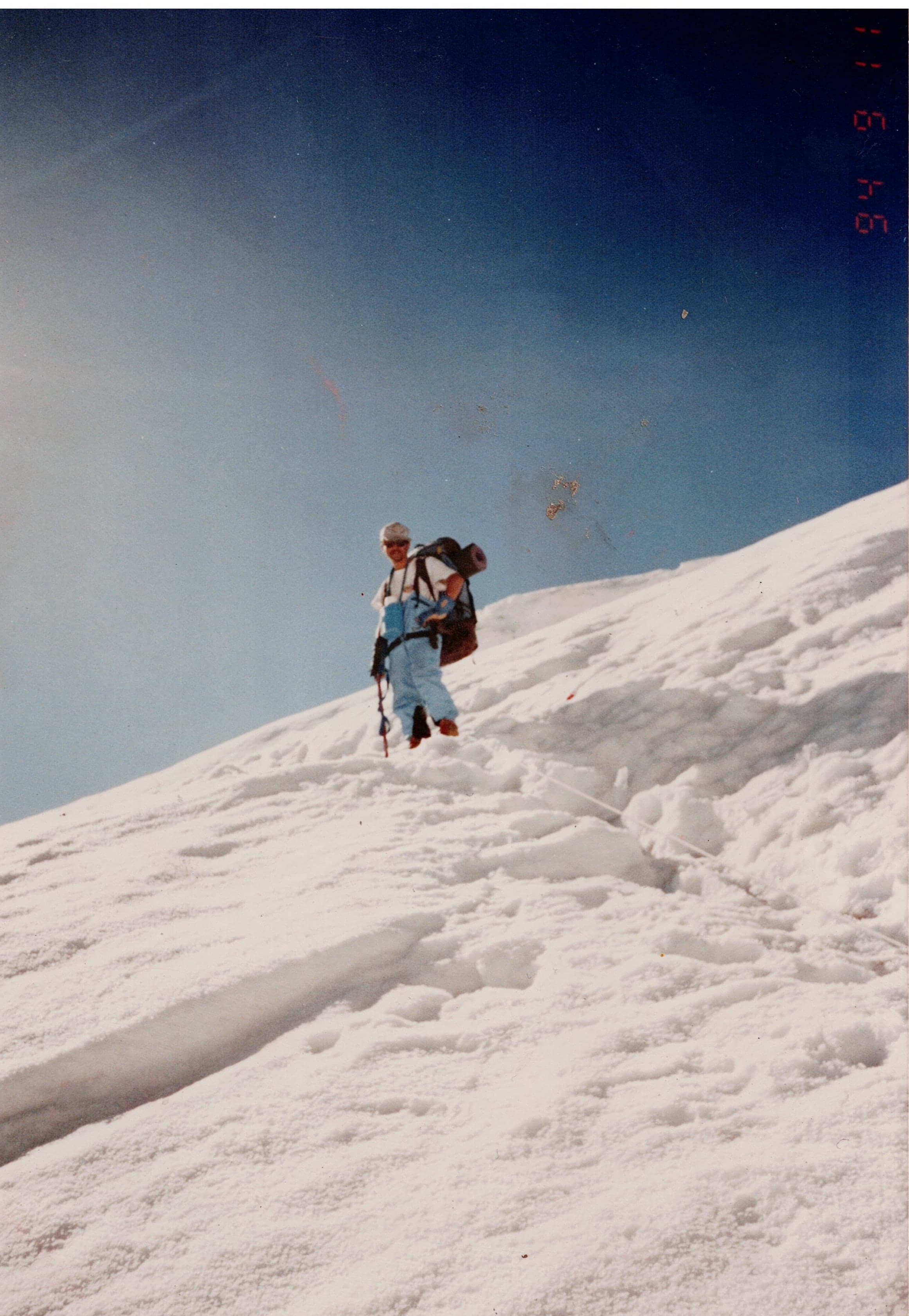 A climber with a large rucksack on a steep snow slope under a deep blue sky