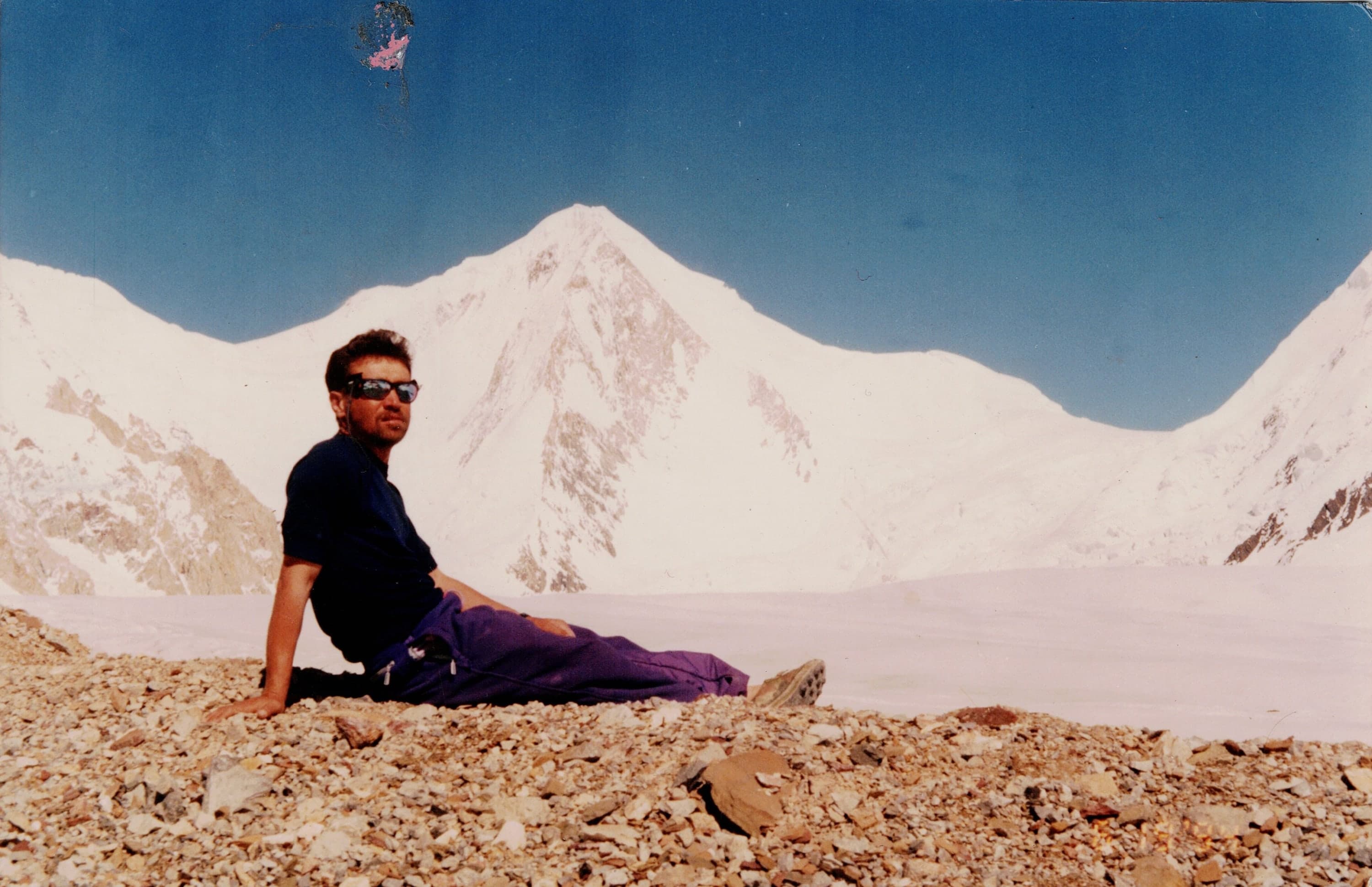 A man in sunglasses reclining on glacier moraine below a large snow peak