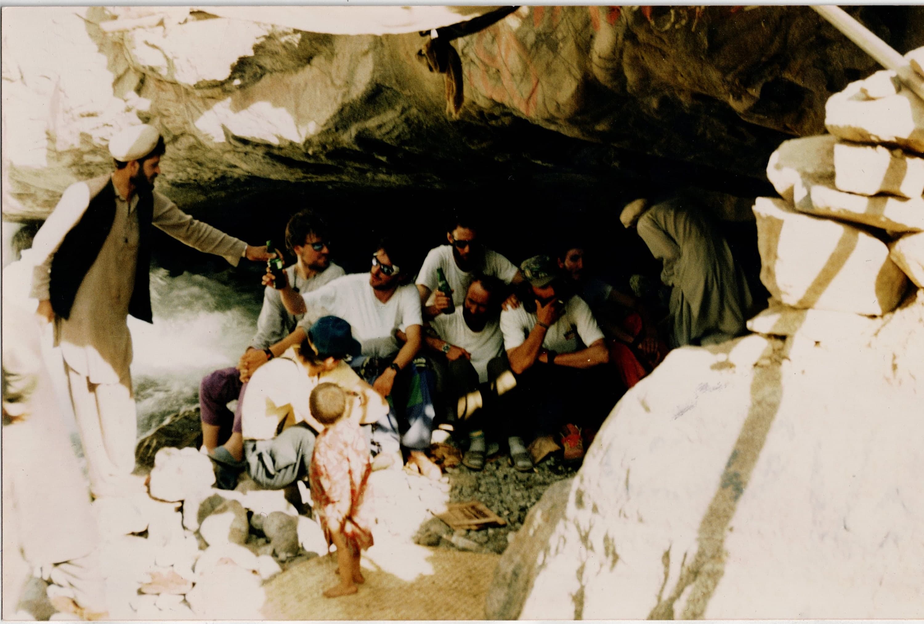Trekkers sitting under a large rock overhang with local men and a child on a Karakoram approach trail