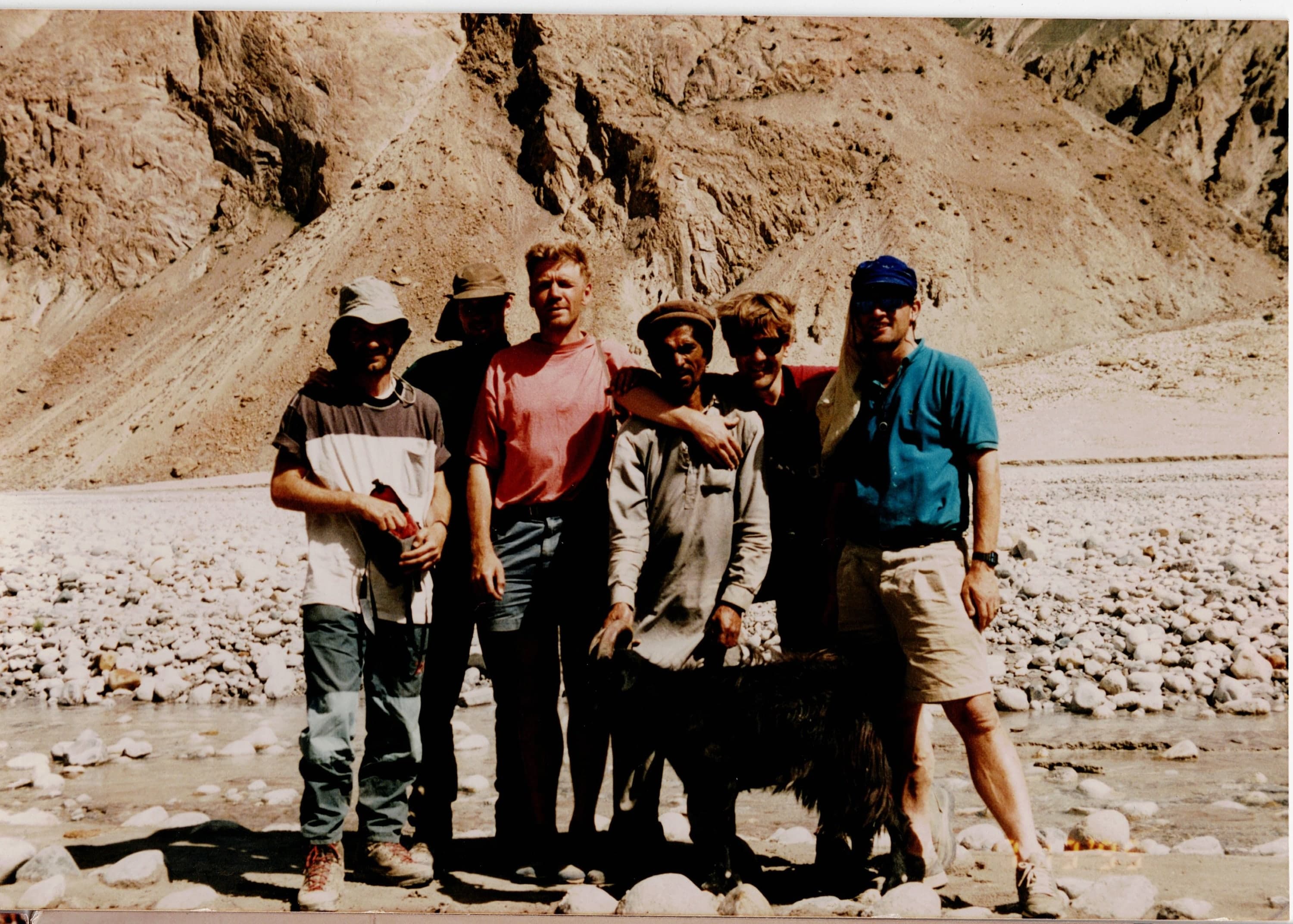 Six trekkers and a goat standing on a stony river bed below dry Karakoram mountainsides